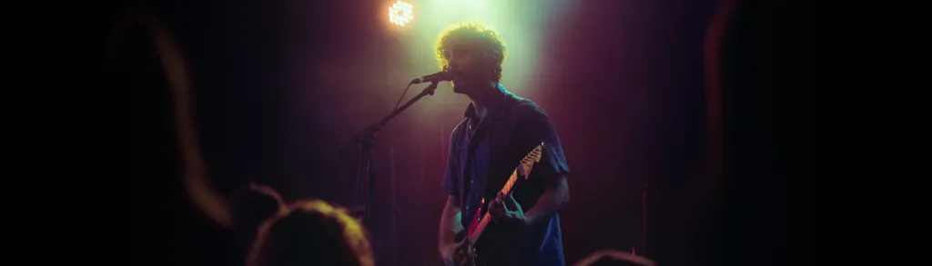 Musician with guitar performing on stage under colorful stage lights, silhouetted against bright yellow and pink lighting.