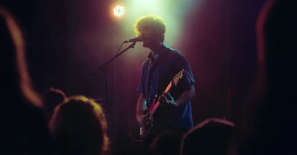 Musiker mit Gitarre auf dunkler Bühne, helles Bühnenlicht im Hintergrund, Publikumssilhouetten im Vordergrund.