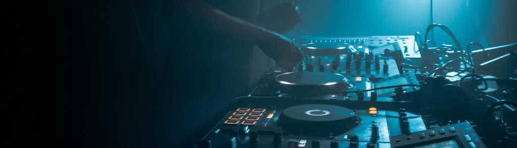 DJ mixing console with turntables, sliders, and control panels illuminated by blue stage lighting in dark venue.