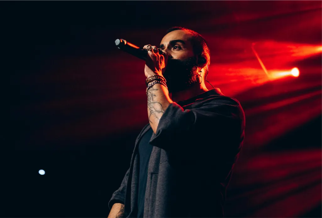 Cantante con mascarilla negra sostiene micrófono, lleva camiseta negra de manga larga, iluminación roja intensa de fondo.