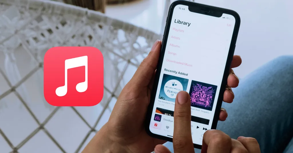 Hand holding iPhone displaying Apple Music library with album covers. Red music note app icon visible in foreground on tiled surface.
