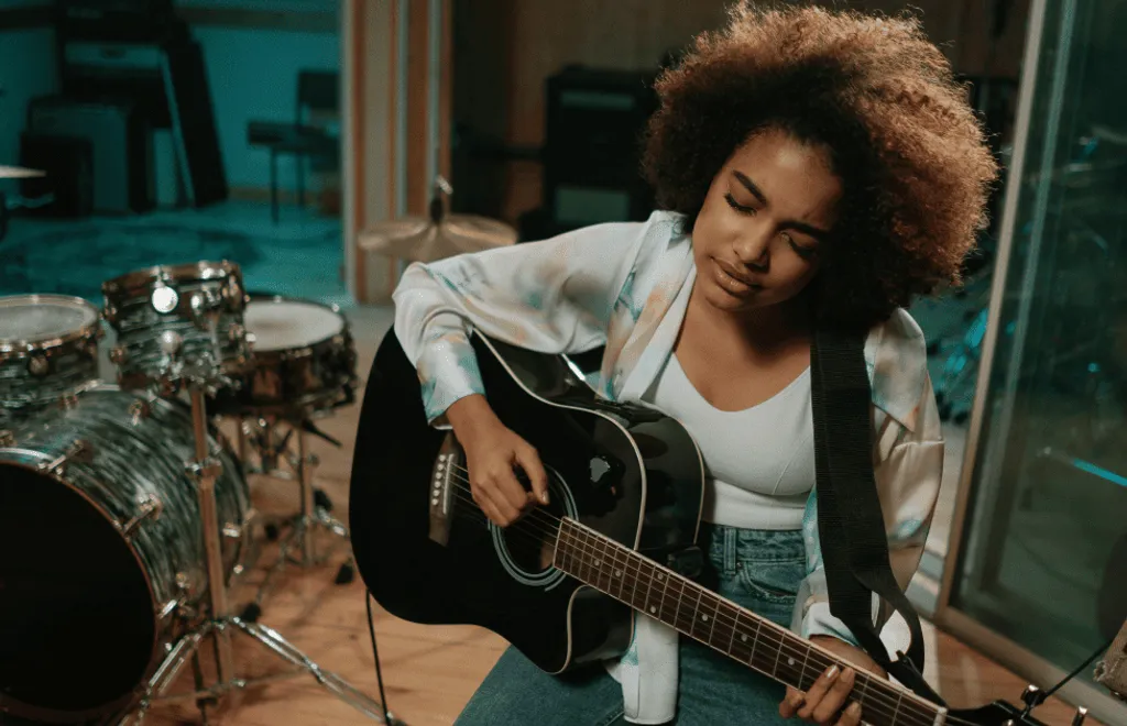 Frau mit lockigem Haar spielt schwarze Akustikgitarre in Musikstudio, Schlagzeug im Hintergrund, trägt weiße Jacke und Jeans.