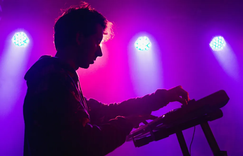 Silhouetted musician at keyboard with bright purple and pink stage lighting, white spotlights creating dramatic backlighting.