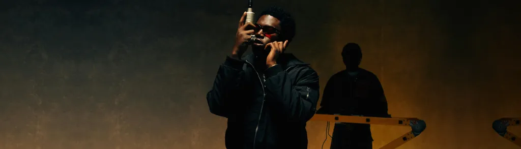 Man in dark clothing drinking from bottle on dimly lit stage with silhouetted figure and microphone stands in background.