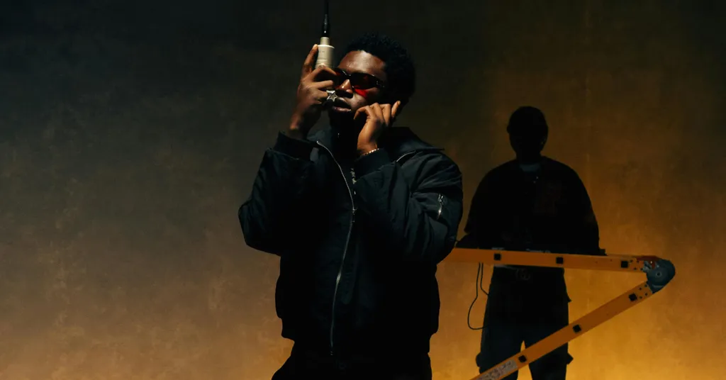 Man in dark jacket holding microphone, another person silhouetted behind podium, warm orange and dark green lighting.