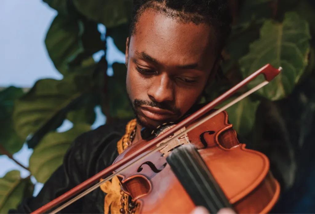 Homme aux yeux fermés jouant du violon, feuillage vert en arrière-plan, lumière naturelle douce.