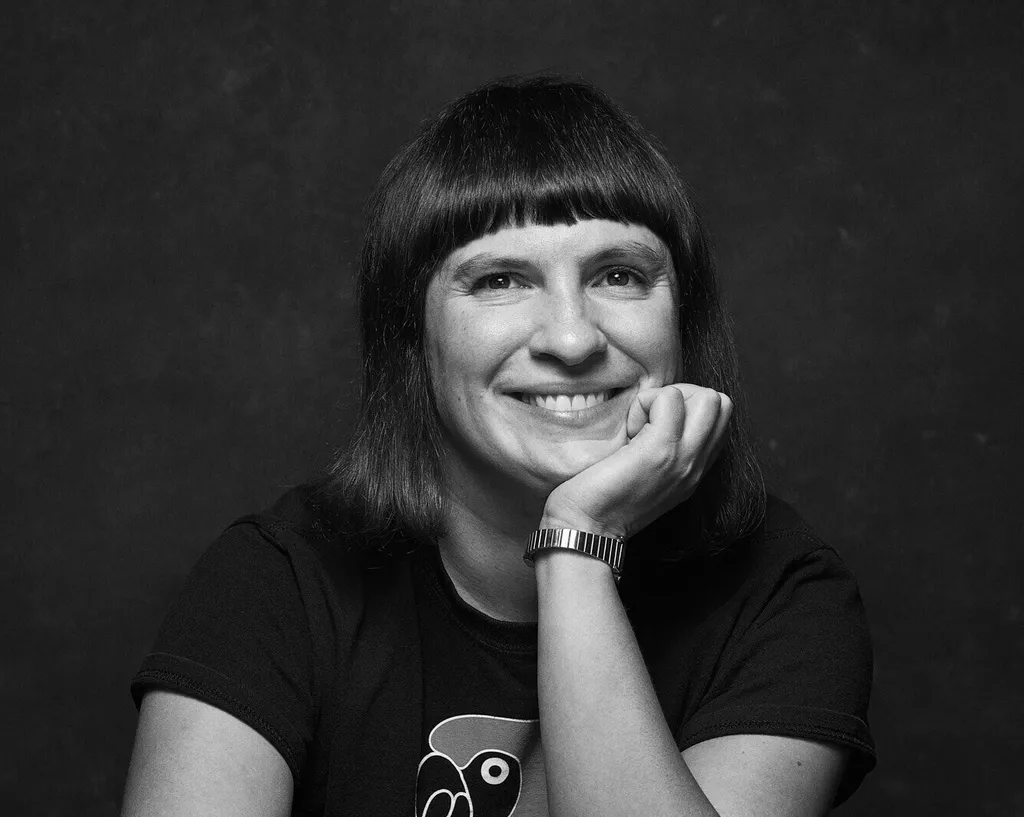 A smiling woman with short, dark hair wearing a black shirt against a dark background.