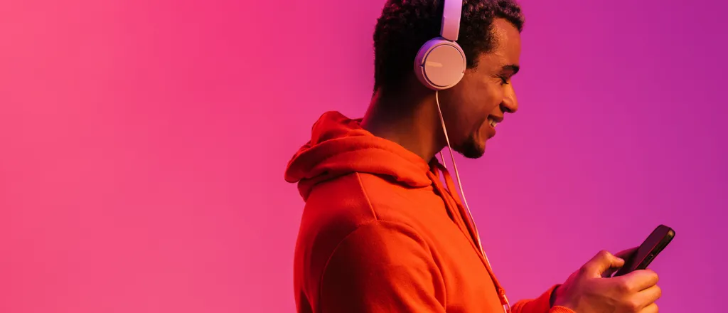 A person wearing white headphones and an orange hoodie, smiling while looking at their smartphone. The background is a gradient of pink to purple.