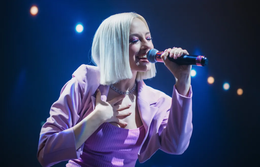 Blonde Frau mit kurzem Bob singt in Mikrofon, trägt lila Outfit, blaue Bühnenbeleuchtung im Hintergrund.