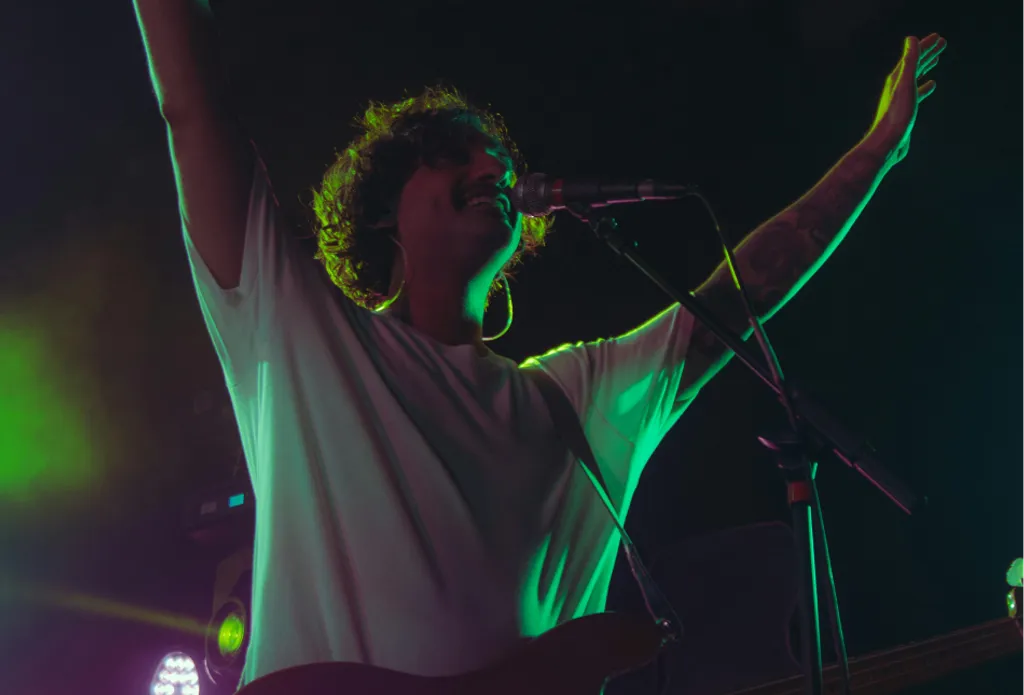 Homme en scène, debout avec les bras levés, chantant dans un microphone. Lumières vertes et bleues éclairant la scène.