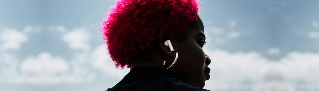 Silhouette di profilo con capelli ricci rosa brillante contro cielo nuvoloso grigio-blu, figura scura in controluce.