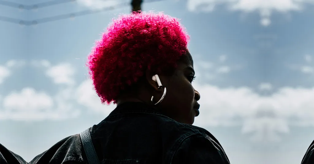 Persona con cabello afro rosa brillante vista de perfil contra cielo azul con nubes blancas, llevando chaqueta oscura.