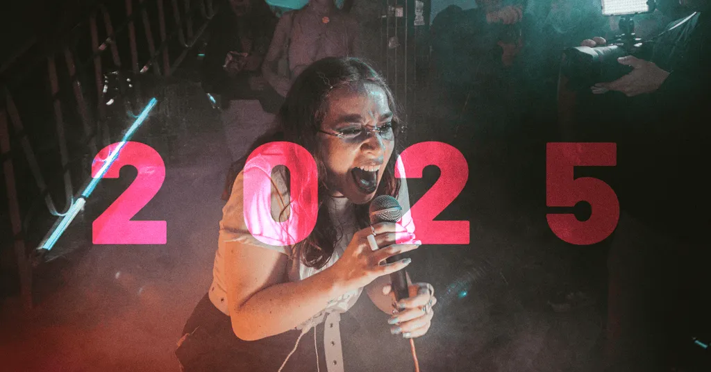 Homme avec microphone devant chiffres "2025" rouges illuminés, éclairage coloré rose et vert, arrière-plan sombre.