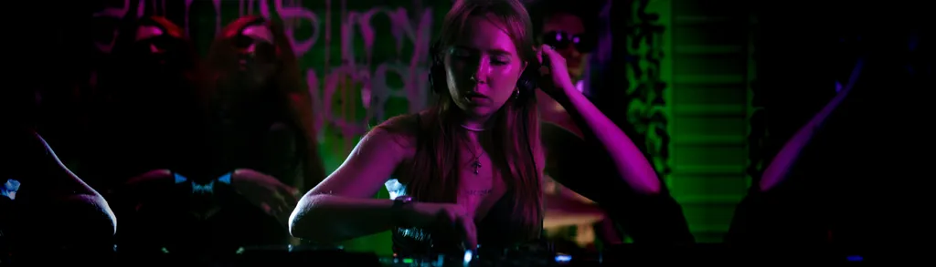 Woman with long hair in dark setting with bright pink and green neon lighting creating colorful shadows and highlights across the scene.