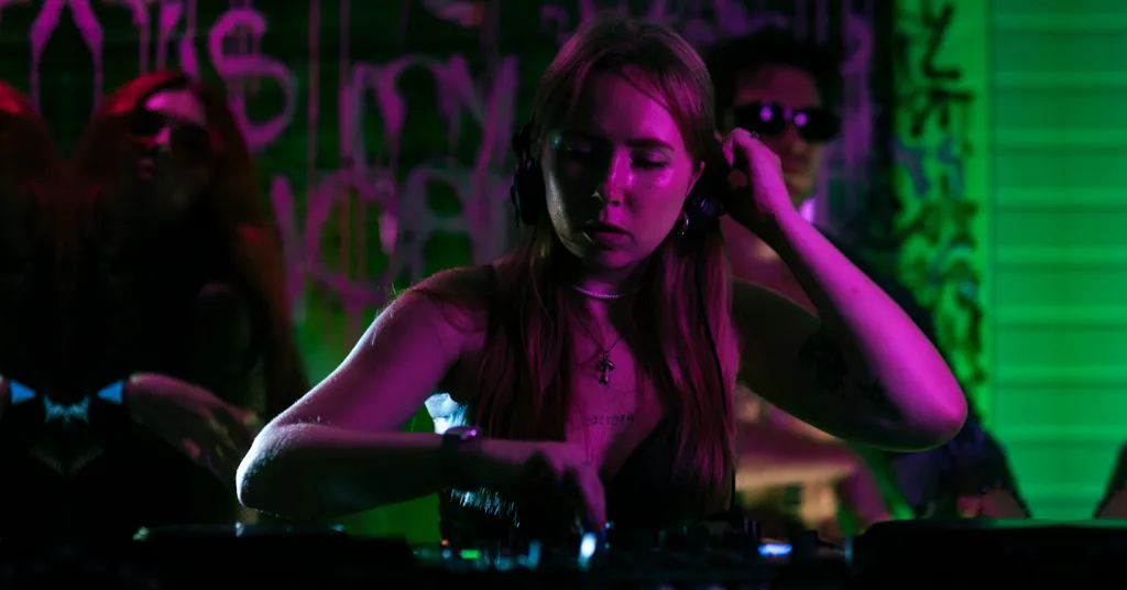 Woman with long dark hair at DJ equipment, illuminated by purple and green stage lighting against dark background with graffiti.