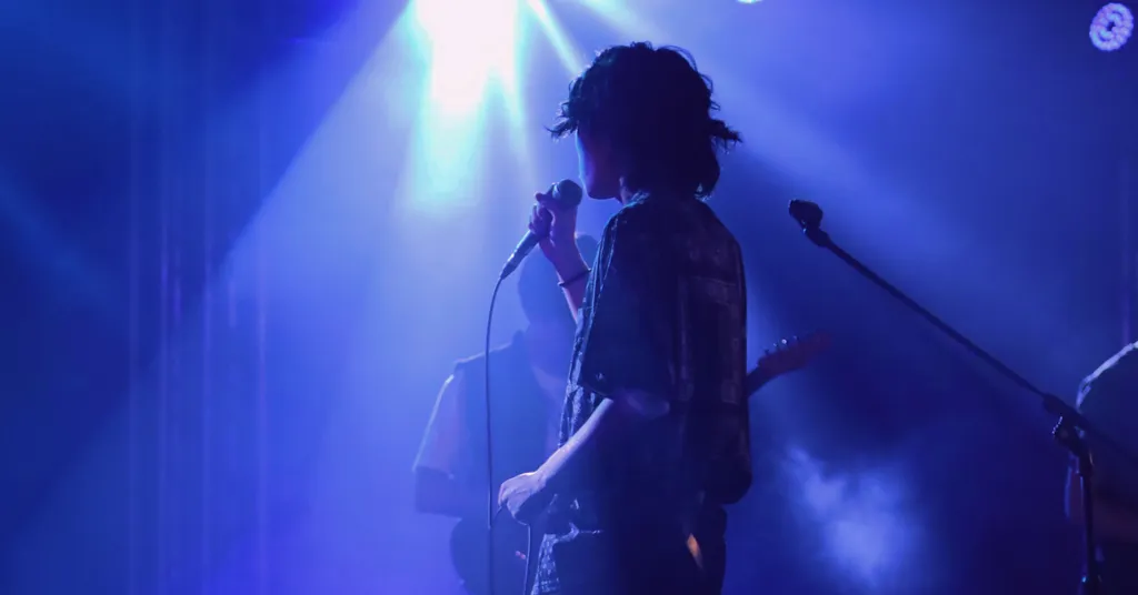 Sänger-Silhouette vor blauem Bühnenlicht, hält Mikrofon, dramatische Beleuchtung mit weißen Strahlen von oben.