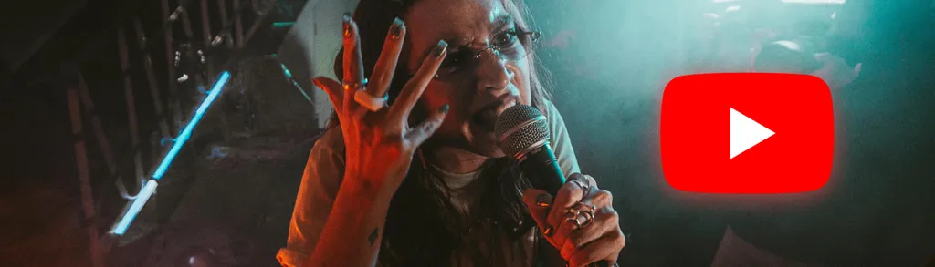 Hombre con cabello largo extendiendo la mano hacia la cámara, iluminación azul y roja, logo de YouTube en esquina superior derecha.