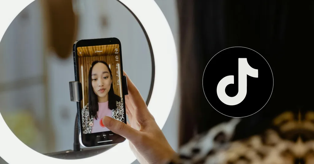 Hand holding smartphone showing woman on screen, white ring light behind phone, TikTok logo visible on right side.