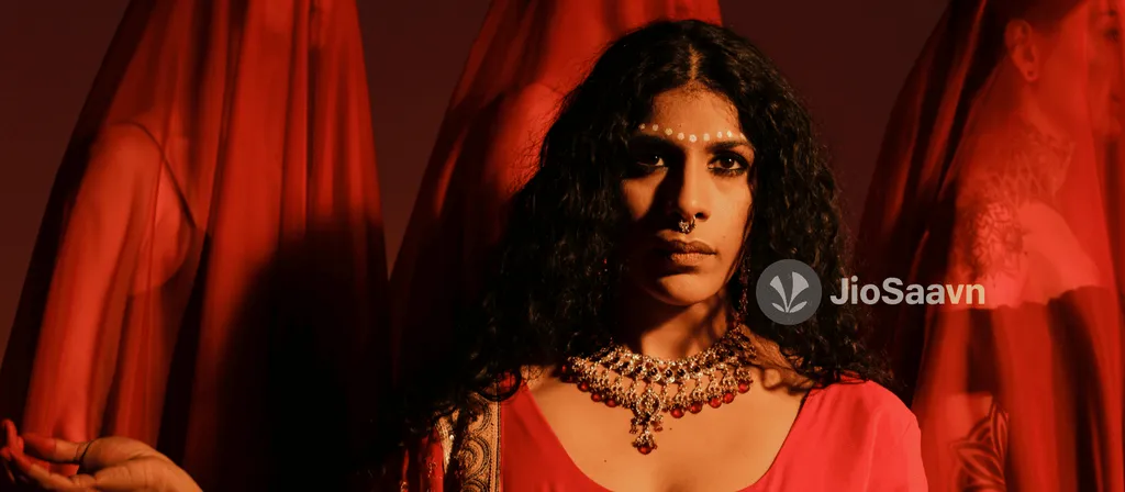 Woman with long dark curly hair wearing ornate gold jewelry and red clothing against red draped fabric background.