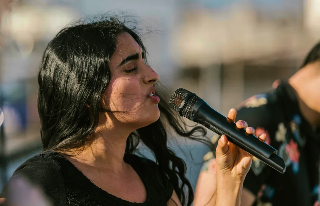 Woman with long dark hair singing into microphone, eyes closed, blurred colorful background with warm lighting.