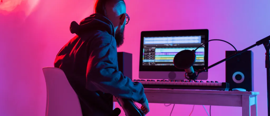 Person sitting at a computer desk in a colorful, neon-lit room. Computer monitor, keyboard, and other equipment visible.