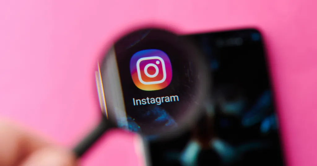 Magnifying glass hovering over Instagram app icon on smartphone screen against bright pink background.