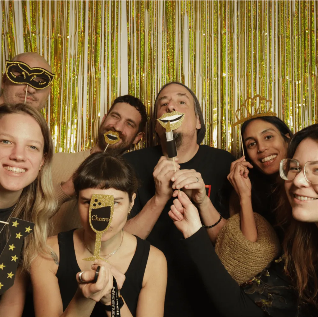 Group of people holding masks and drinks, smiling at the camera against a glittery gold backdrop.