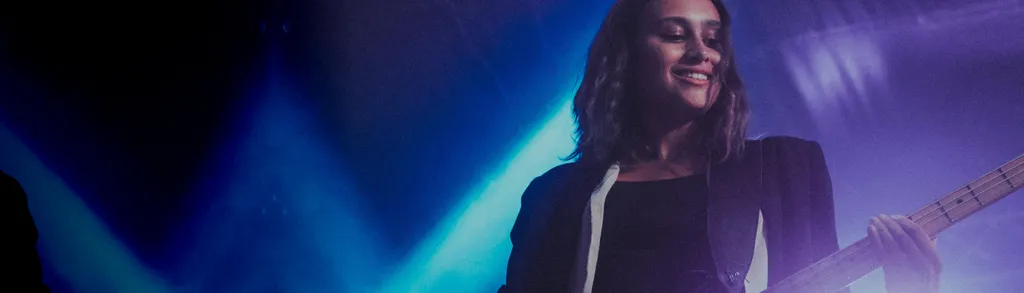 Smiling woman with long dark hair holding guitar against blue and purple stage lighting background.