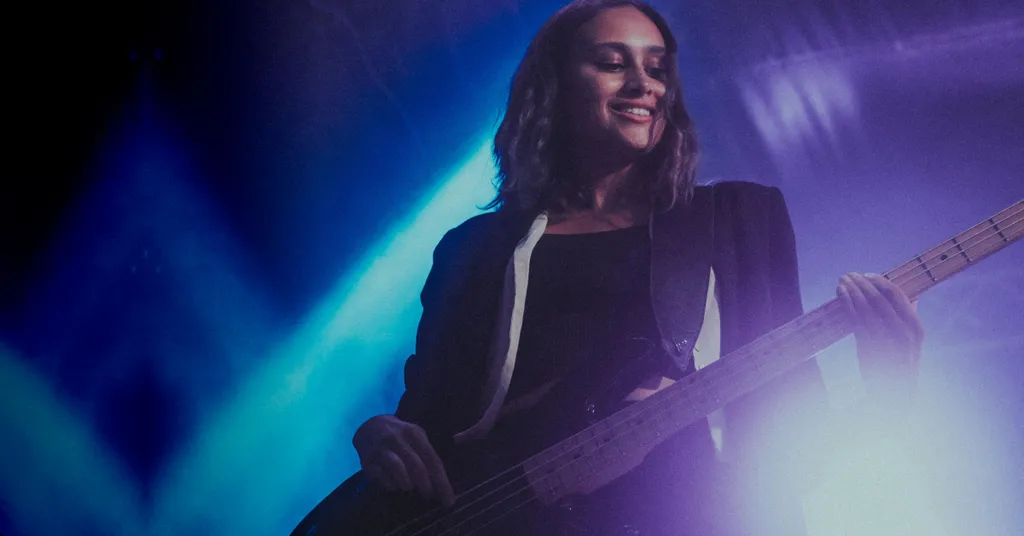 Frau mit langen dunklen Haaren hält Gitarre, lächelt. Blaues und violettes Bühnenlicht, Nebel im Hintergrund.