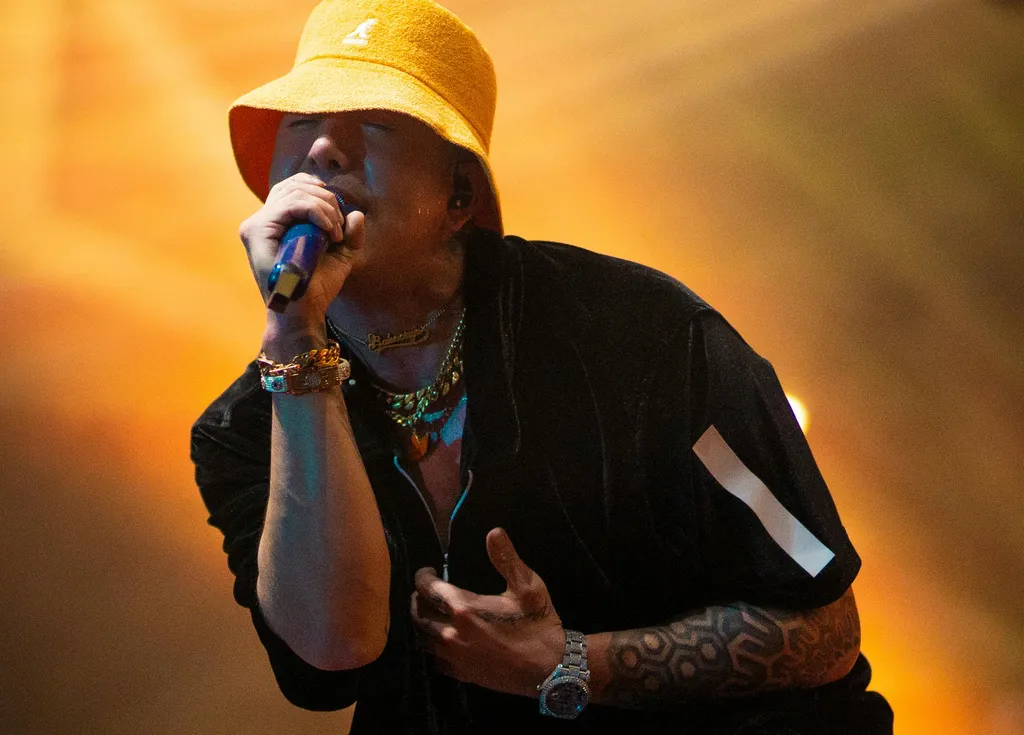 A person wearing a yellow hat and sunglasses performing on stage, with bright orange and yellow lighting in the background.
