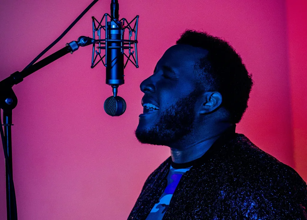 A man with a beard and curly hair, dressed in a dark jacket, singing into a blue microphone in front of a pink and blue background.