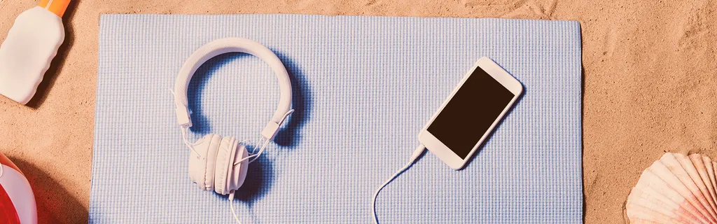 Kopfhörer und iPhone liegen auf Strandtuch und Sand Sonnencreme Muschel