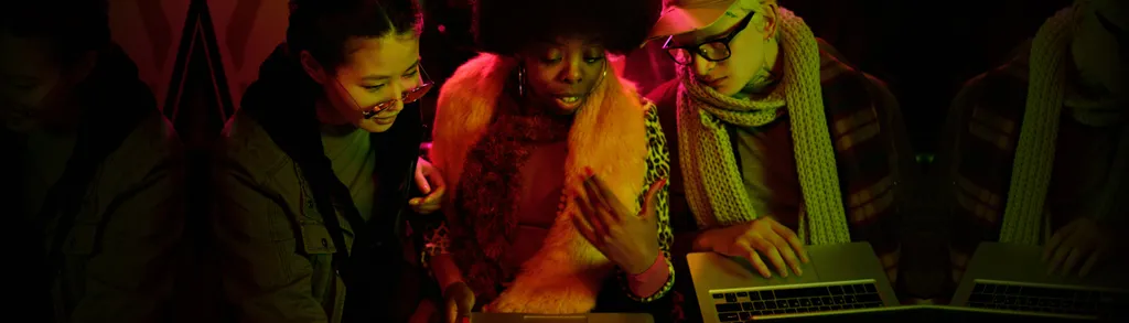 Group of people in dimly lit space with red and green lighting, faces partially visible in shadows, warm orange and yellow tones on clothing.