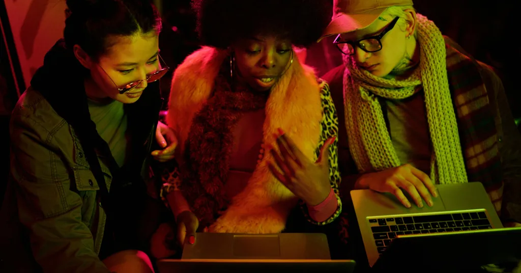 Three women gathered around laptop computer, illuminated by red and pink lighting against dark background.