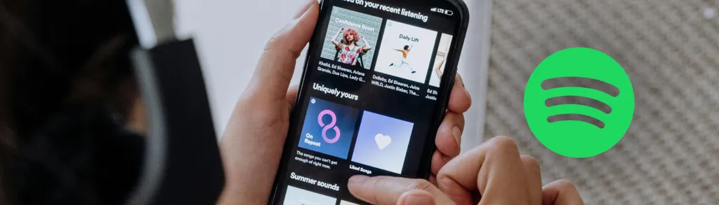 Hand holding smartphone displaying Spotify app interface with album covers and playlists. Green Spotify logo visible in corner.