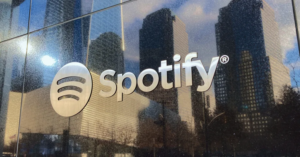 Logo de Spotify en ventana de vidrio con reflejo de edificios urbanos y cielo azul con nubes blancas.