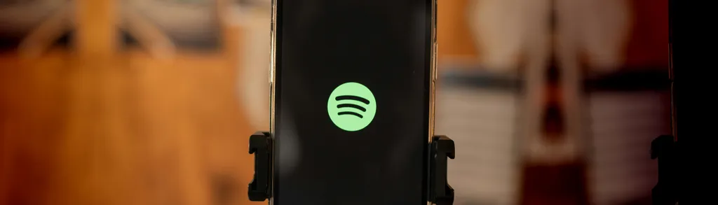 Smartphone in black stand displaying green Spotify logo on dark screen, with blurred wooden interior background.