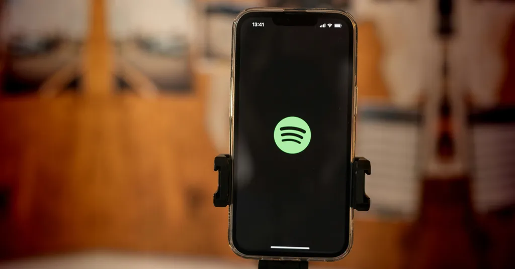 Teléfono móvil en soporte mostrando logo verde de Spotify en pantalla negra, fondo borroso con tonos cálidos.