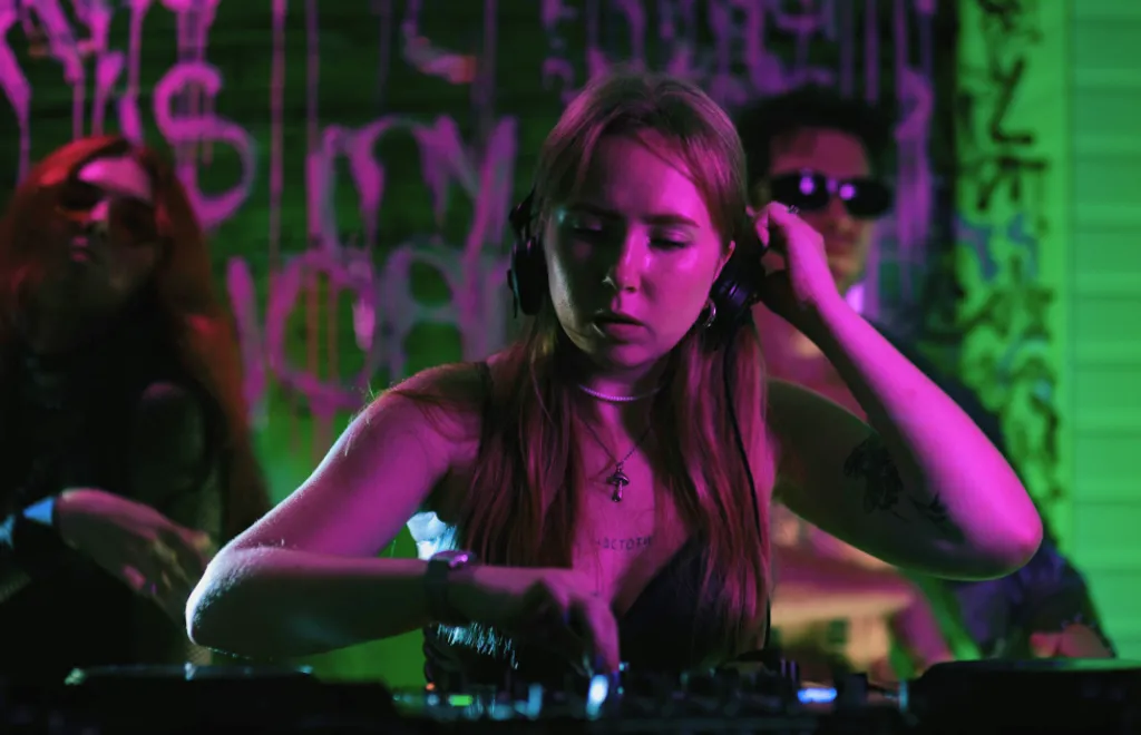 DJ-Frau mit Kopfhörern in violettem und grünem Neonlicht, Graffiti-Wand im Hintergrund, Hand am Kopf, Mischpult vor ihr.