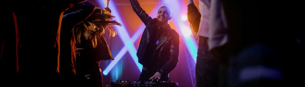 DJ em silhueta atrás de equipamentos com luzes roxas e azuis, multidão com braços erguidos em primeiro plano escuro.