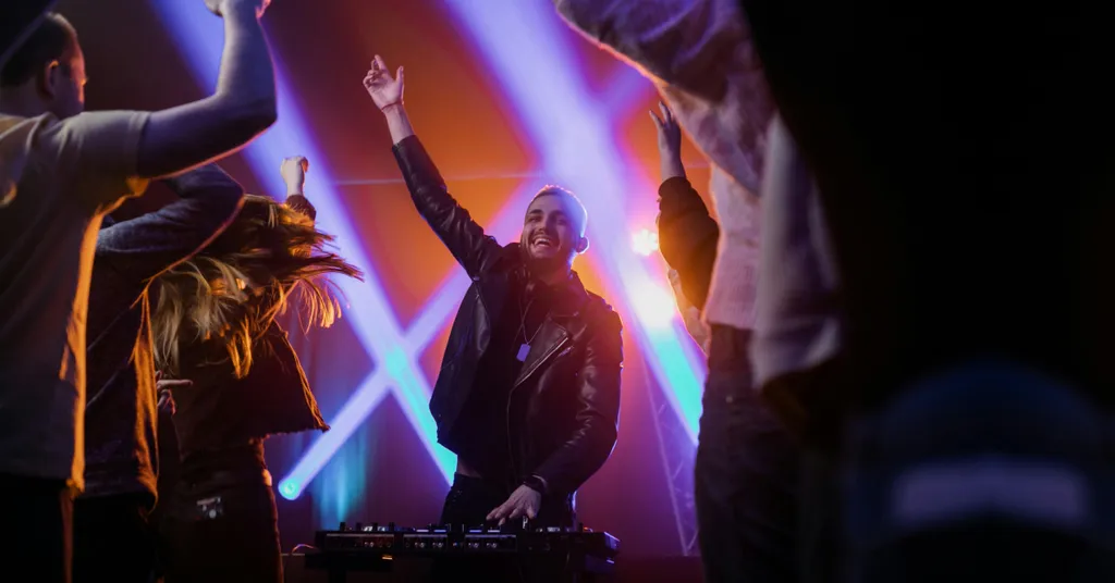 DJ e dançarinos com braços levantados no palco, iluminados por feixes de luz coloridos roxos, azuis e laranjas em show noturno.