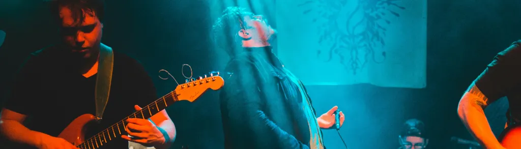 Tre musicisti con chitarre sotto luci colorate blu e arancioni durante un concerto al chiuso.