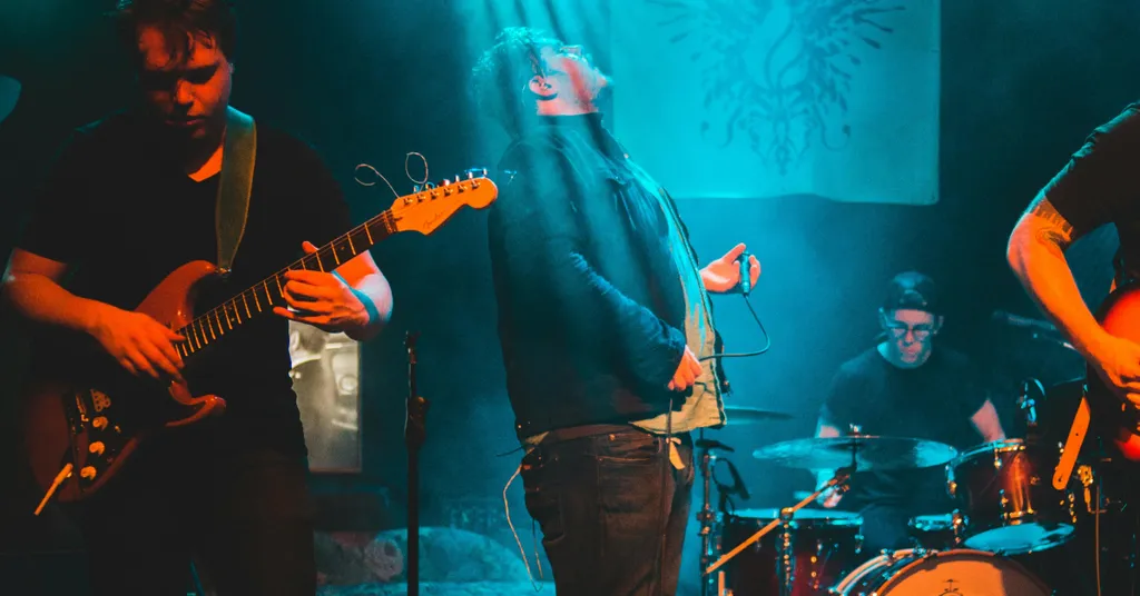 Banda tocando en escenario con iluminación azul y naranja. Guitarrista a la izquierda, cantante al centro, baterista al fondo.