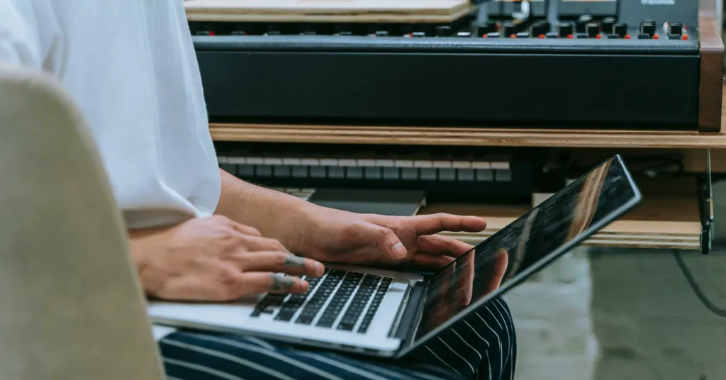Person tippt auf Laptop-Tastatur, Musikmischpult mit Reglern im Hintergrund, Homestudio-Setup.
