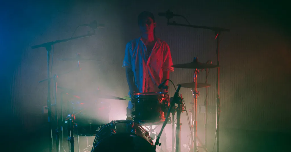 Verschwommene Aufnahme eines Schlagzeugers in rotem und blauem Licht, dunkle Schatten im Hintergrund, körnige Textur.