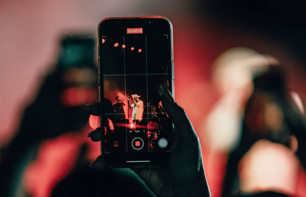 Hands holding smartphone recording a video of a performer on stage, with red stage lighting and blurred crowd in the background.