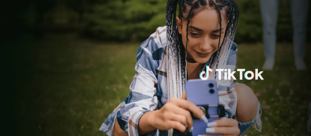 Person with braided hair in striped shirt holding smartphone with TikTok logo visible on screen, green, nature-related background.