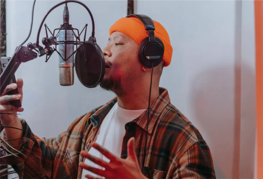 Mann mit oranger Mütze und Kopfhörern singt in Studiomikrofon, trägt kariertes Hemd über weißem T-Shirt.