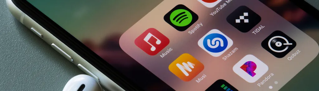 iPhone screen showing app icons including Spotify, TextSoap, Shazam, and others, with white earbuds visible at bottom left corner.