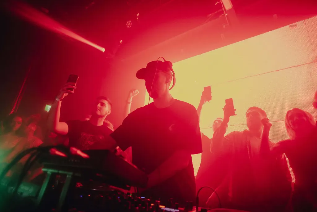 DJ performing at mixing console with crowd dancing, bathed in bright red and pink stage lighting with visible light beams overhead.